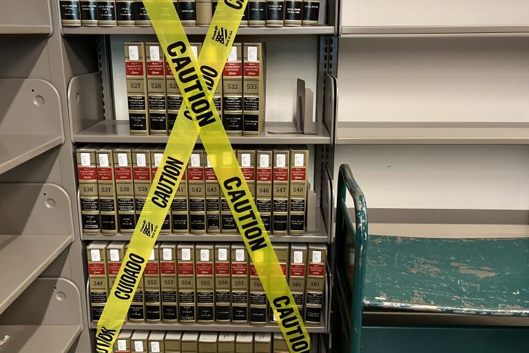Photo of a shelf of books with yellow "Caution" tape crossed in front.