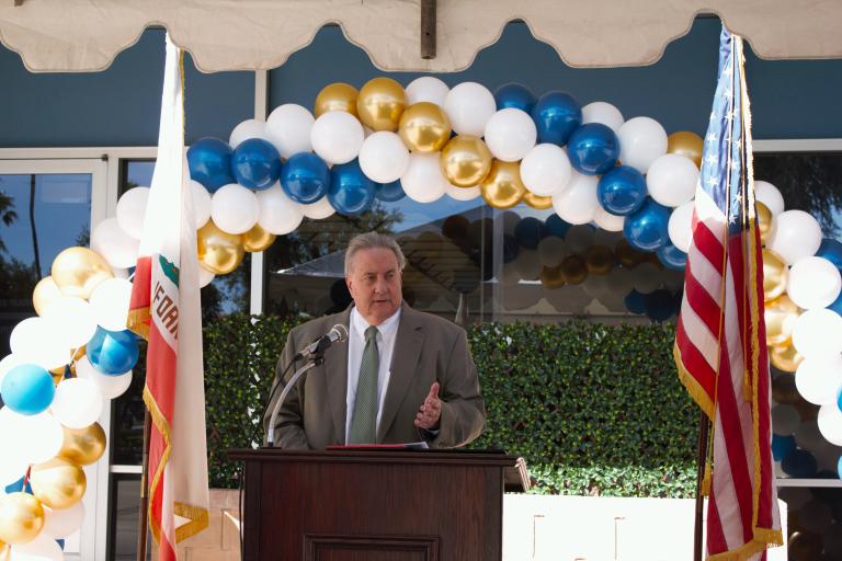 Board President John Boyd delivering remarks from a podium in front of a balloon arch outside the Palm Springs Law Library.