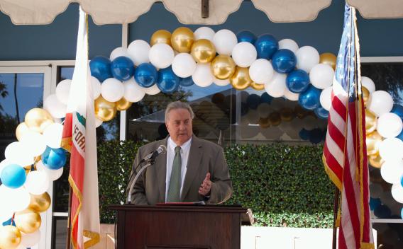 Board President John Boyd delivering remarks from a podium in front of a balloon arch outside the Palm Springs Law Library.