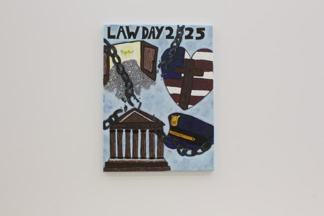 The top of Dreier’s artwork says, “Law Day 2025”. On the top left of the piece, there are people gathered around two large open doors where the inside says, “together.” To the right of this is the US flag in the shape of a heart with a brown cross in the middle reading, “religion.” Below the heart is a blue police hat with a plaque that says “safety.” To the left of the hat and below the doors, there is a brown structure of columns with the words “Justice” on the front. At the forefront, there are chains sn