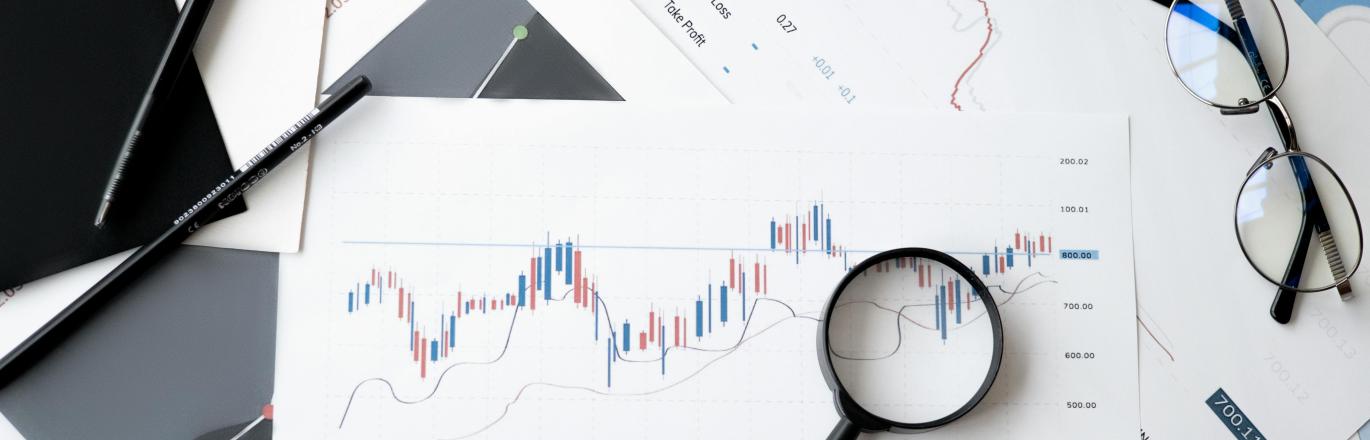 A desk covered in printed stock charts, pens, a laptop, a magnifying glass, and a pair of glasses.