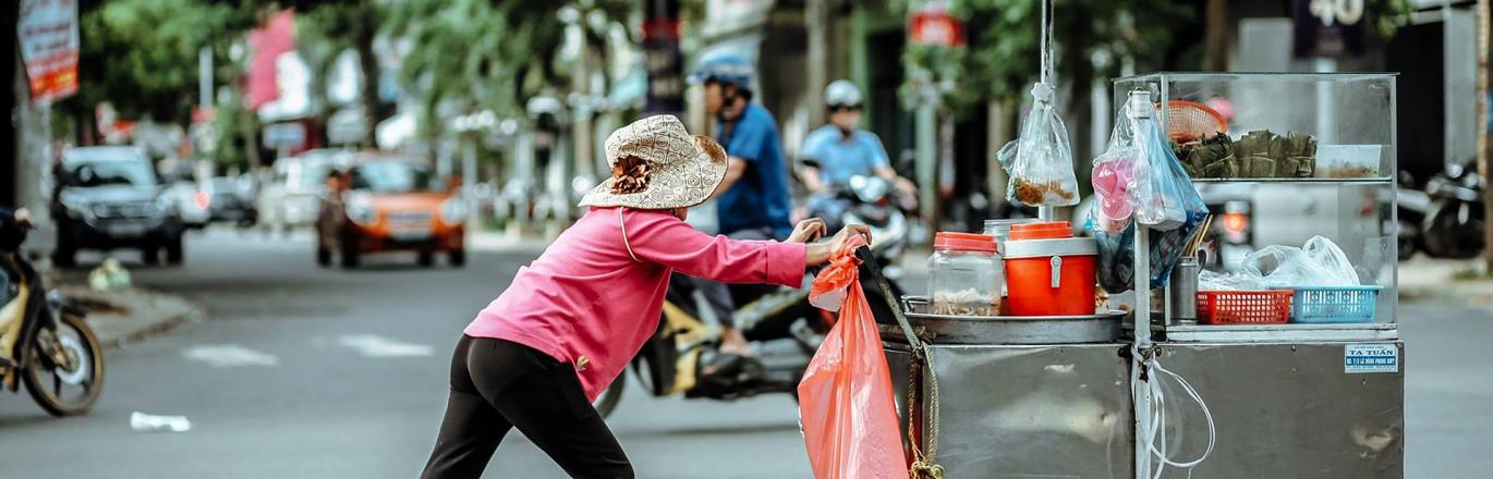 female appearing street vendor pushing a metal cart with an orange and green shade umbrella in the middle of a street