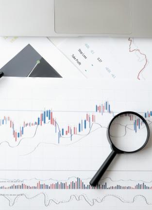 A desk covered in printed stock charts, pens, a laptop, a magnifying glass, and a pair of glasses.