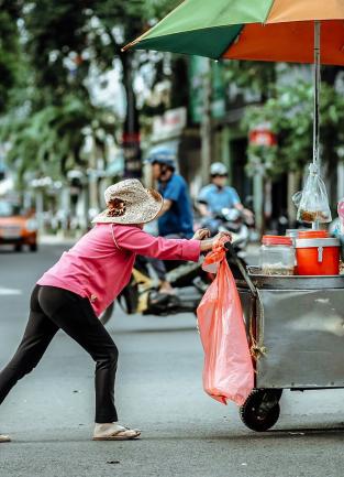 female appearing street vendor pushing a metal cart with an orange and green shade umbrella in the middle of a street